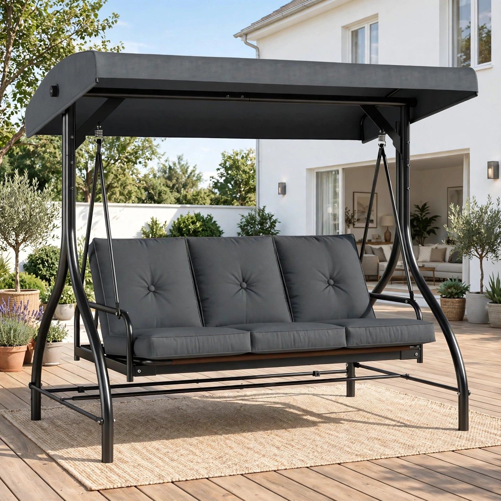 Charcoal gray 3-seater patio swing chair with adjustable canopy on sunny wooden deck with garden and house backdrop
